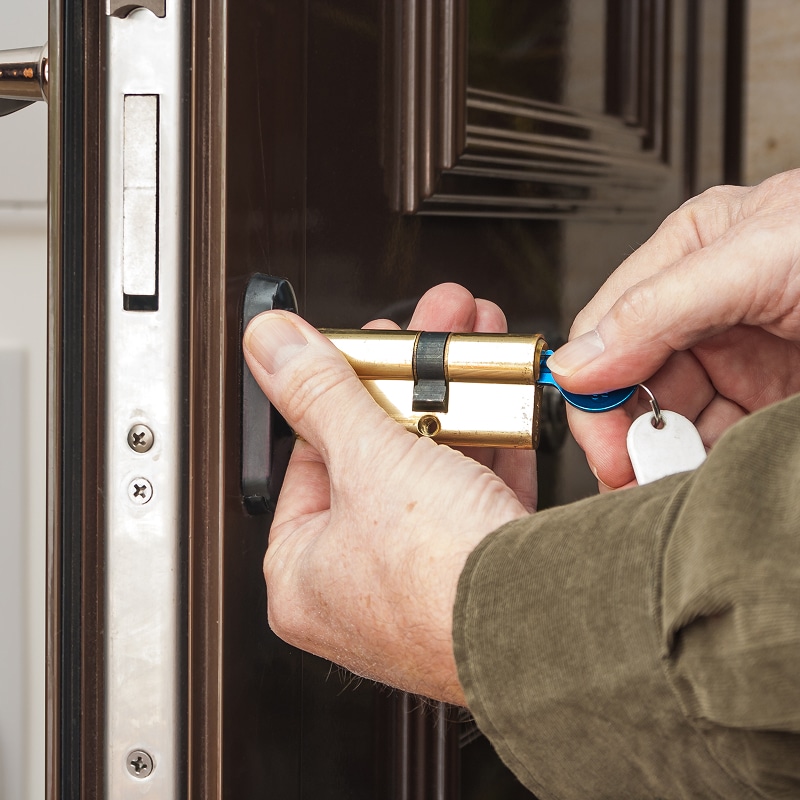 Mains manipulant un cylindre de serrure en laiton, insérant une clé bleue près d'une porte marron. Installation ou test de sécurité.