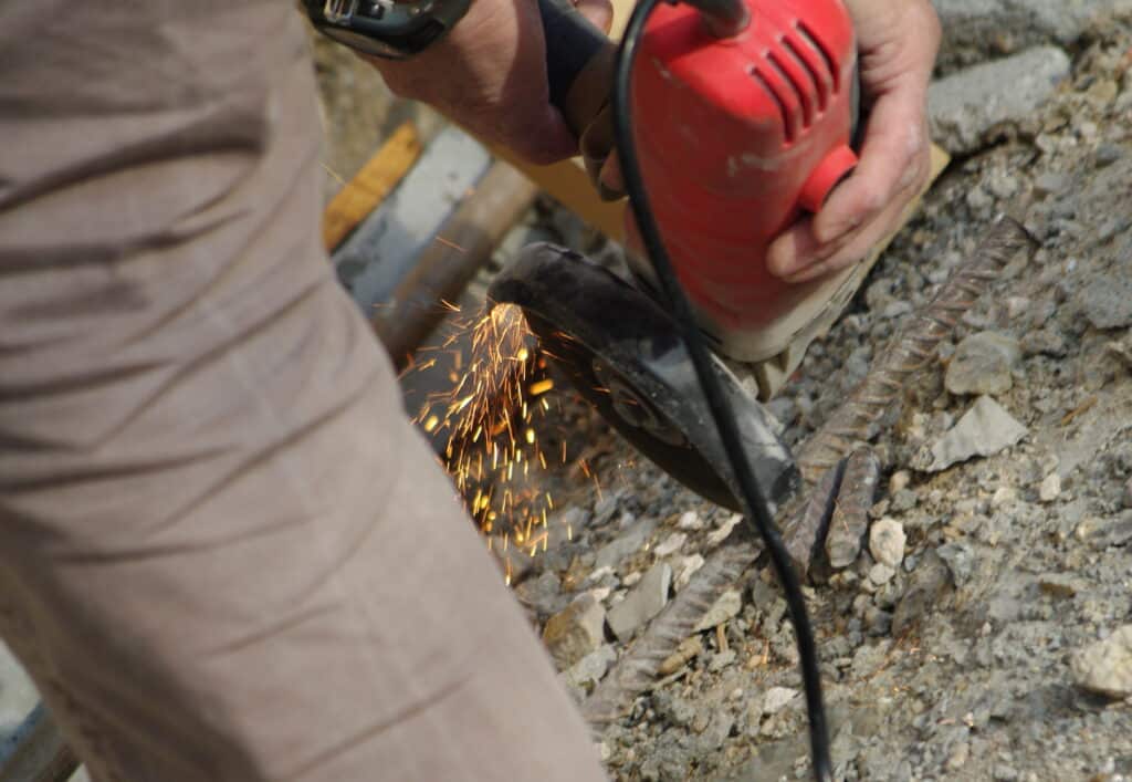 Travailleur utilisant une meuleuse d'angle rouge pour couper une barre d'armature, générant un jet d'étincelles intenses sur le chantier.
