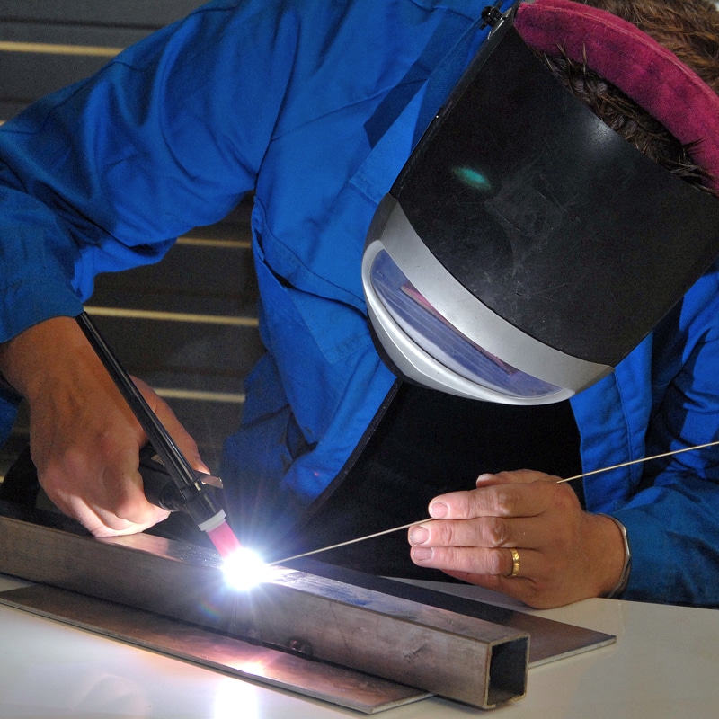 Personne en combinaison bleue utilisant un poste TIG pour souder du métal, masque de soudeur baissé et étincelles vives.