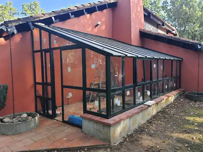 Véranda ou serre moderne en cours d'installation, structure noire vitrée sur muret de briques, adossée à un mur terracotta.