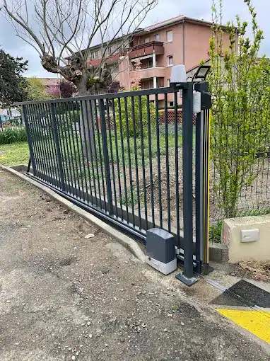Portail coulissant motorisé gris anthracite : Installation moderne Portail coulissant gris anthracite motorisé avec feu clignotant et panneau solaire, installé devant un immeuble résidentiel et un grand arbre.
